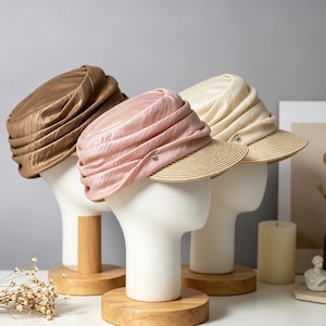 May include: Three hats on mannequin heads. The hats are in shades of gold, pink, and cream, each with a pleated fabric crown and a straw brim. The hats are displayed on wooden stands, with a neutral background.