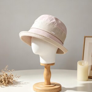 Puede incluir: Un sombrero de pescador rosa claro y beige con una peque&ntilde;a etiqueta met&aacute;lica. El sombrero se exhibe sobre una cabeza de maniqu&iacute; blanca sobre un soporte de madera. El sombrero tiene un ala ancha y una corona estructurada. El fondo incluye un cuadro enmarcado, una vela y un jarr&oacute;n.