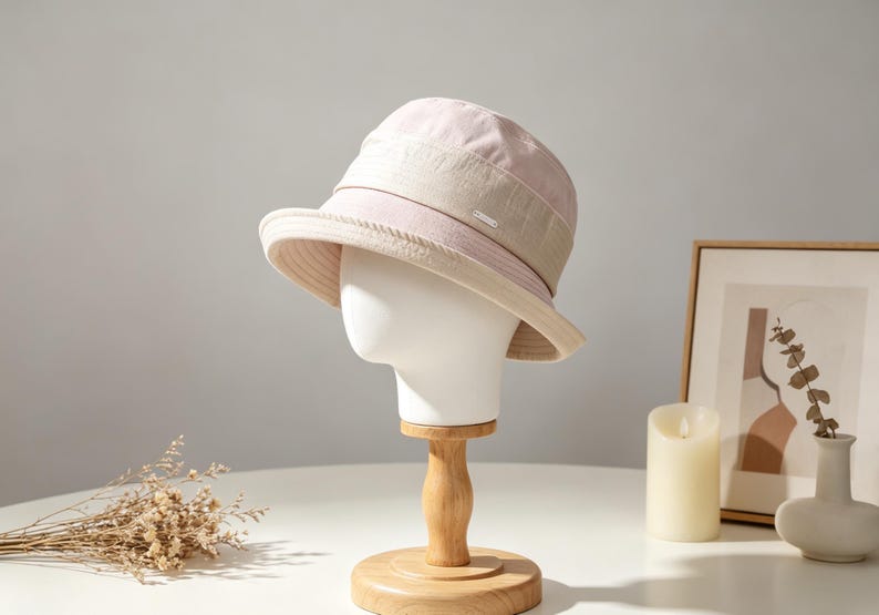Puede incluir: Un sombrero de pescador rosa claro y beige con una peque&ntilde;a etiqueta met&aacute;lica. El sombrero se exhibe sobre una cabeza de maniqu&iacute; blanca sobre un soporte de madera. El sombrero tiene un ala ancha y una corona estructurada. El fondo incluye un cuadro enmarcado, una vela y un jarr&oacute;n.