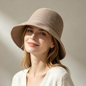 May include: A beige sun hat with a wide brim and a structured crown. The hat has a textured appearance and is worn by a person with blonde hair and a white top. The hat is designed to provide shade and protection from the sun.
