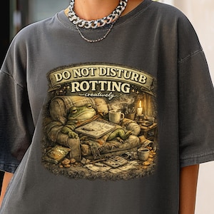 May include: Dark grey t-shirt featuring a whimsical illustration of a frog relaxing on a couch, reading a book, with the text "DO NOT DISTURB ROTTING creatively". The frog is surrounded by books, a candle, and coffee cups.