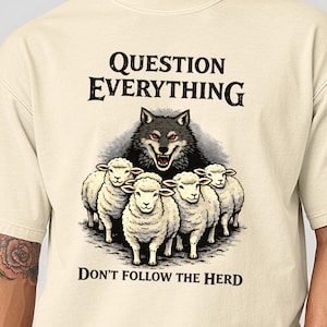 May include: Cream-colored t-shirt featuring a wolf with red eyes looming over a group of sheep. The shirt has the text "QUESTION EVERYTHING" above the wolf and "DON'T FOLLOW THE HERD" below the sheep.