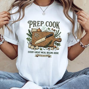 May include: A white t-shirt featuring a graphic of a wooden cutting board with a knife, onions, garlic, mushrooms, and herbs. The text reads PREP COOK, EVERY GREAT MEAL BEGINS HERE, and CRAFTED WITH PRIDE. The design is centered on the chest area.
