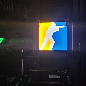 May include: A computer case with a glowing logo featuring a white silhouette aiming a gun, set against a yellow and blue background. The text "GEFORCE RTX" is visible on a component inside the case.