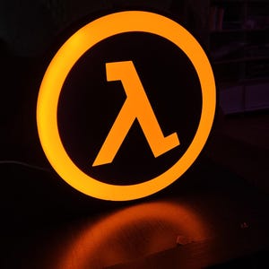 Peut inclure: Un panneau lumineux orange et noir représentant le symbole lambda, une lettre grecque souvent utilisée en science et en mathématiques. Le panneau est circulaire avec un fond noir et une bordure et un symbole orange vif. Le panneau est éclairé, dégageant une lueur chaleureuse.