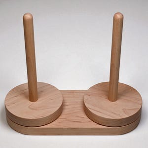 May include: A light-colored wooden yarn holder with two vertical dowels. The base is rectangular with rounded corners, and the dowels are centered on circular platforms. The wood grain is visible.