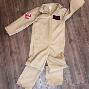 May include: A beige long-sleeve jumpsuit featuring a central gold zipper, a collar, and two vertical pockets. A circular ghost emblem patch is on the right sleeve, and a rectangular black patch with the red text ABELE is on the left chest.