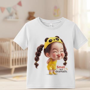 May include: White t-shirt featuring a cartoon illustration of a girl with pigtails, wearing a yellow panda hat and outfit. The text "Small but dramatic" is printed on the shirt. The shirt is designed for children.