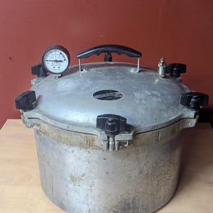 May include: A silver pressure canner with a black handle and black knobs. The canner has a gauge on top and is sitting on a wooden surface. The canner is used for preserving food.