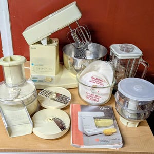 May include: An off-white Oster Kitchen Center appliance set including a stand mixer with a metal bowl, a blender pitcher, a food processor attachment with two metal slicing discs, a small glass bowl with a caution label, and an instruction manual.