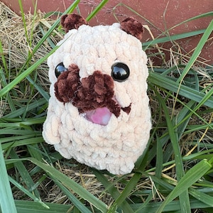 May include: A handmade, plush pug dog toy. The toy is made of light beige yarn with dark brown accents for the ears and snout. It has large, black, shiny eyes and a pink tongue. The pug is sitting in green grass.