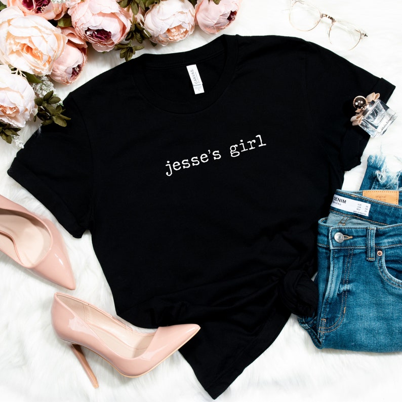 May include: Black t-shirt with white text that reads "Jesse's girl" on a white background. The shirt is laid flat with a pair of pink high heels, a pair of blue jeans, and a small bottle of perfume.
