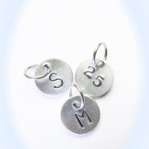 May include: Three silver metal charms with jump rings. One charm has the letter "S" engraved on it, another has the number "25" engraved on it, and the third has the letter "M" engraved on it.
