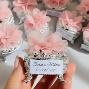 Può includere: Una collezione di piccole scatole argentate ornate, ognuna sormontata da un delicato fiore di tessuto rosa e una gemma scintillante. Una scatola è tenuta in mano, con i nomi "Tomas & Melanie" e la data "12/12/2025".