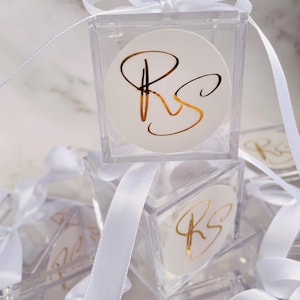 May include: Clear acrylic favor boxes with white ribbon and bows. Each box features a white circular sticker with gold lettering of the initials "RS". The boxes are stacked, creating a visually appealing display.