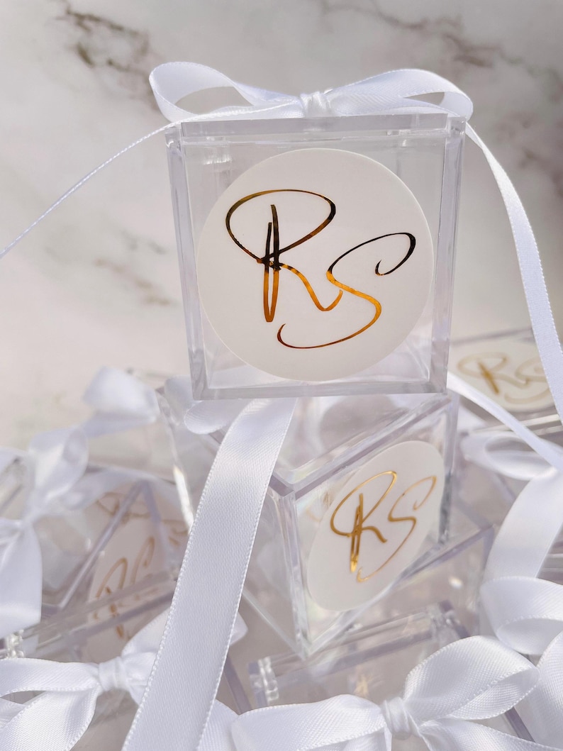 May include: Clear acrylic favor boxes with white ribbon and bows. Each box features a white circular sticker with gold lettering of the initials "RS". The boxes are stacked, creating a visually appealing display.