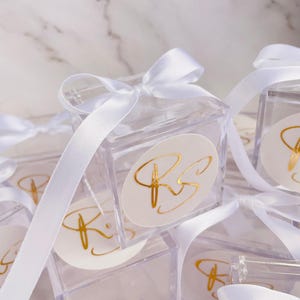 May include: Clear acrylic favor boxes tied with white satin ribbon. Each box features a white oval sticker with gold calligraphy initials. The boxes are arranged on a white marble surface.