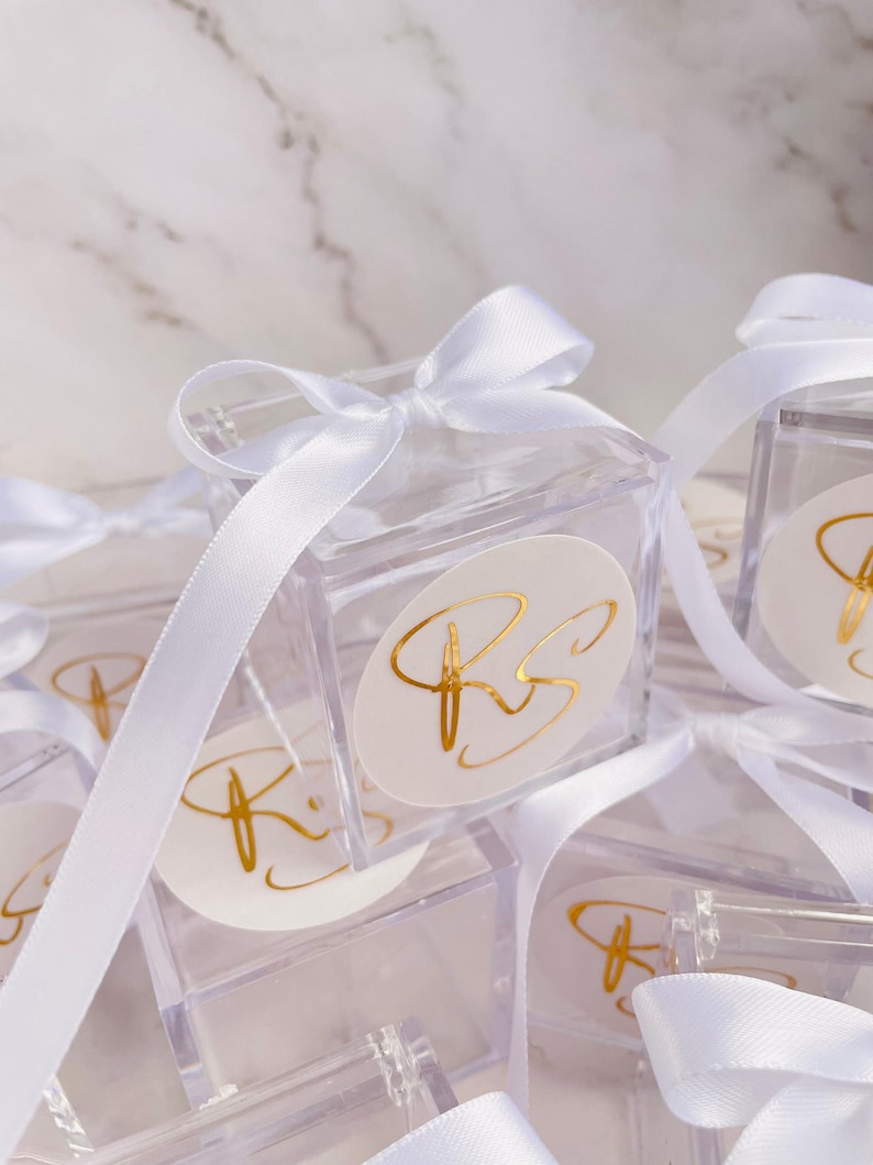 May include: Clear acrylic favor boxes tied with white satin ribbon. Each box features a white oval sticker with gold calligraphy initials. The boxes are arranged on a white marble surface.