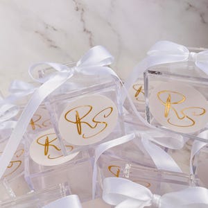 May include: Clear acrylic favor boxes tied with white satin ribbons. Each box features a gold foil sticker with the initials "RS". The boxes are arranged on a white marble surface, creating a clean and elegant presentation.