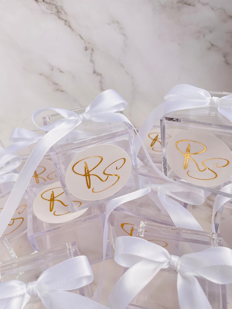 May include: Clear acrylic favor boxes tied with white satin ribbons. Each box features a gold foil sticker with the initials "RS". The boxes are arranged on a white marble surface, creating a clean and elegant presentation.