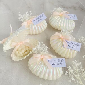 Op de afbeelding: Iriserende schelpvormige gunsten, vastgebonden met perzikkleurige linten en versierd met gipskruid. Elke schelp bevat kleine witte bollen en een label met de tekst "Emanuel & Jeniffer 20.12.2025".