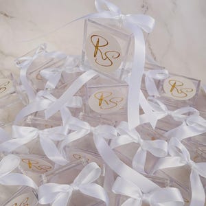 May include: A collection of clear, square boxes with white ribbon bows. Each box has a gold monogram on a white circular sticker. The boxes are arranged on a white marble surface.