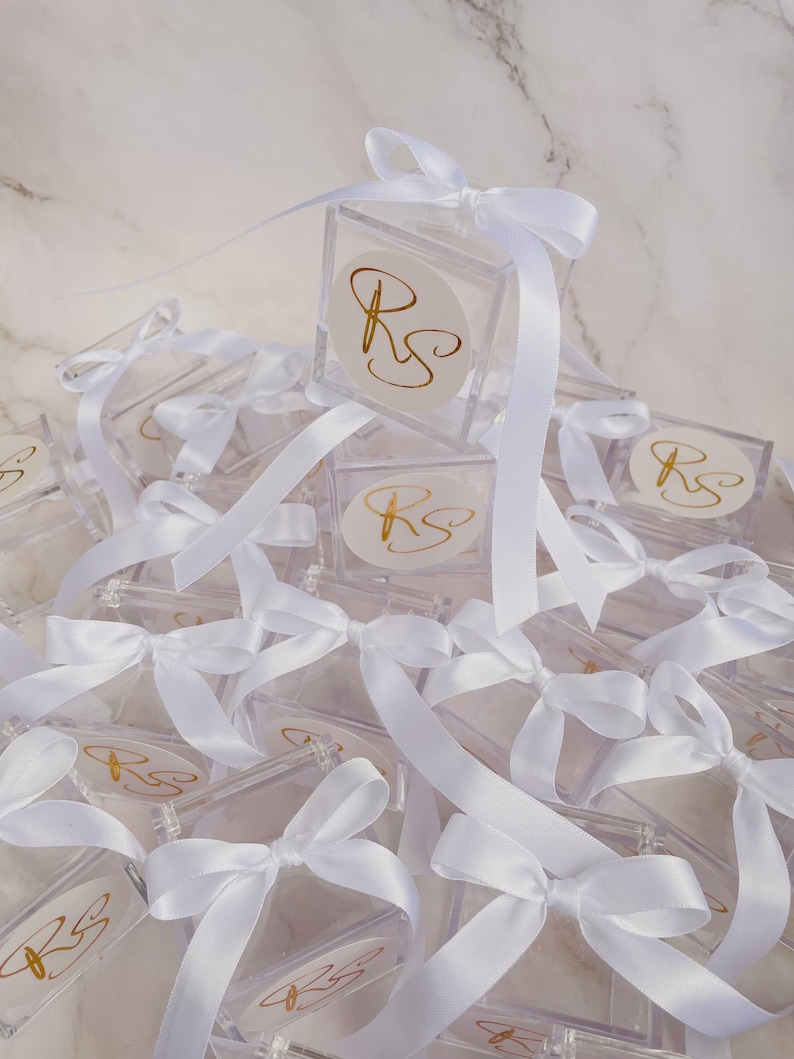 May include: A collection of clear, square boxes with white ribbon bows. Each box has a gold monogram on a white circular sticker. The boxes are arranged on a white marble surface.