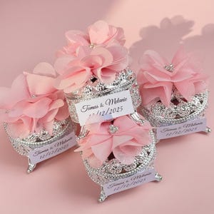 Op de afbeelding: Versierde zilverkleurige sieradendoosjes met roze bloemaccenten en een klein label met de tekst "Tomas & Melanie 12/12/2025". Elk doosje heeft een decoratief ontwerp en is afgewerkt met een roze stoffen bloem.