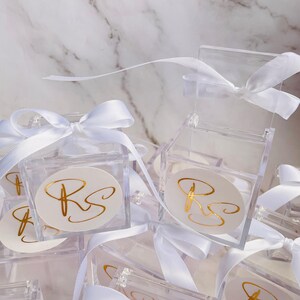 May include: Clear acrylic favor boxes tied with white satin ribbons. Each box features a white oval sticker with gold script initials. The boxes are arranged on a white marble surface.