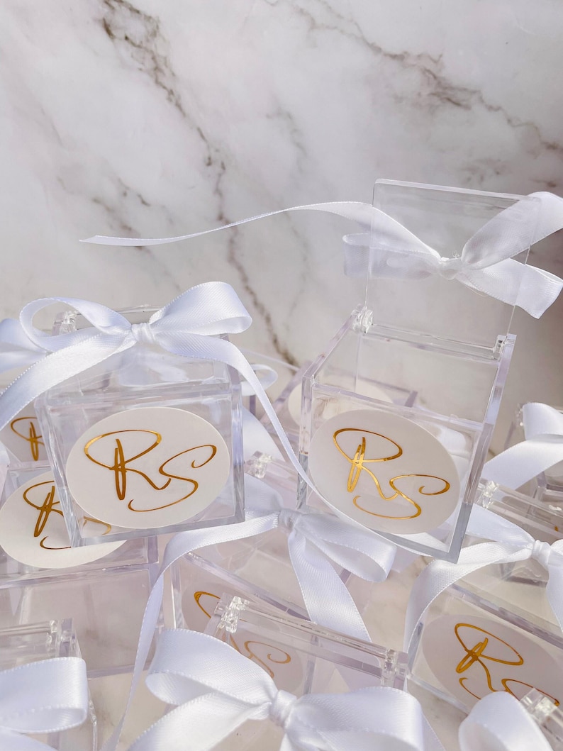 May include: Clear acrylic favor boxes tied with white satin ribbons. Each box features a white oval sticker with gold script initials. The boxes are arranged on a white marble surface.