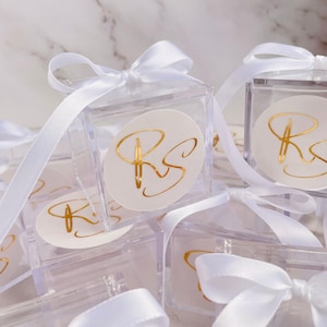 May include: Clear acrylic favour boxes tied with white satin ribbons. Each box has a white circular sticker with gold lettering. The boxes are grouped together, some in focus and others blurred.