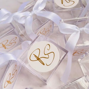 May include: Clear square boxes tied with white satin ribbons. Each box has a white circular label with gold-colored initials "RS". The boxes are arranged on a white marble surface, suggesting a celebratory or gift-giving theme.