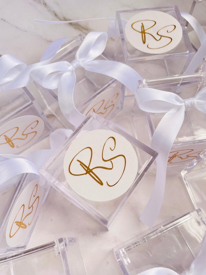May include: Clear square boxes tied with white satin ribbons. Each box has a white circular label with gold-colored initials "RS". The boxes are arranged on a white marble surface, suggesting a celebratory or gift-giving theme.
