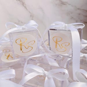 May include: Clear square favor boxes tied with white satin ribbons. Each box features a round sticker with gold script initials. The boxes are arranged on a white marble surface, creating a delicate and elegant presentation.