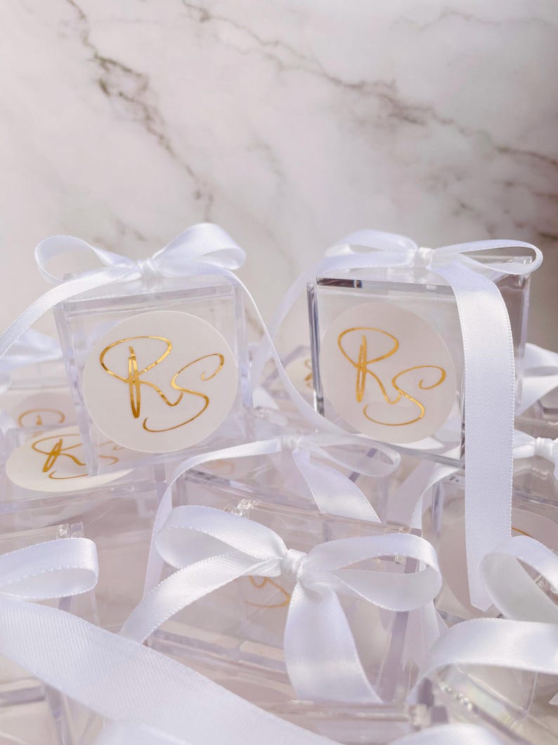 May include: Clear square favor boxes tied with white satin ribbons. Each box features a round sticker with gold script initials. The boxes are arranged on a white marble surface, creating a delicate and elegant presentation.