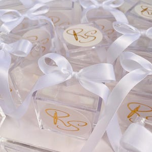 May include: Clear acrylic boxes tied with white satin ribbons and bows. Each box features a gold monogram on a white oval sticker. The boxes are arranged closely together, creating a visually appealing display.