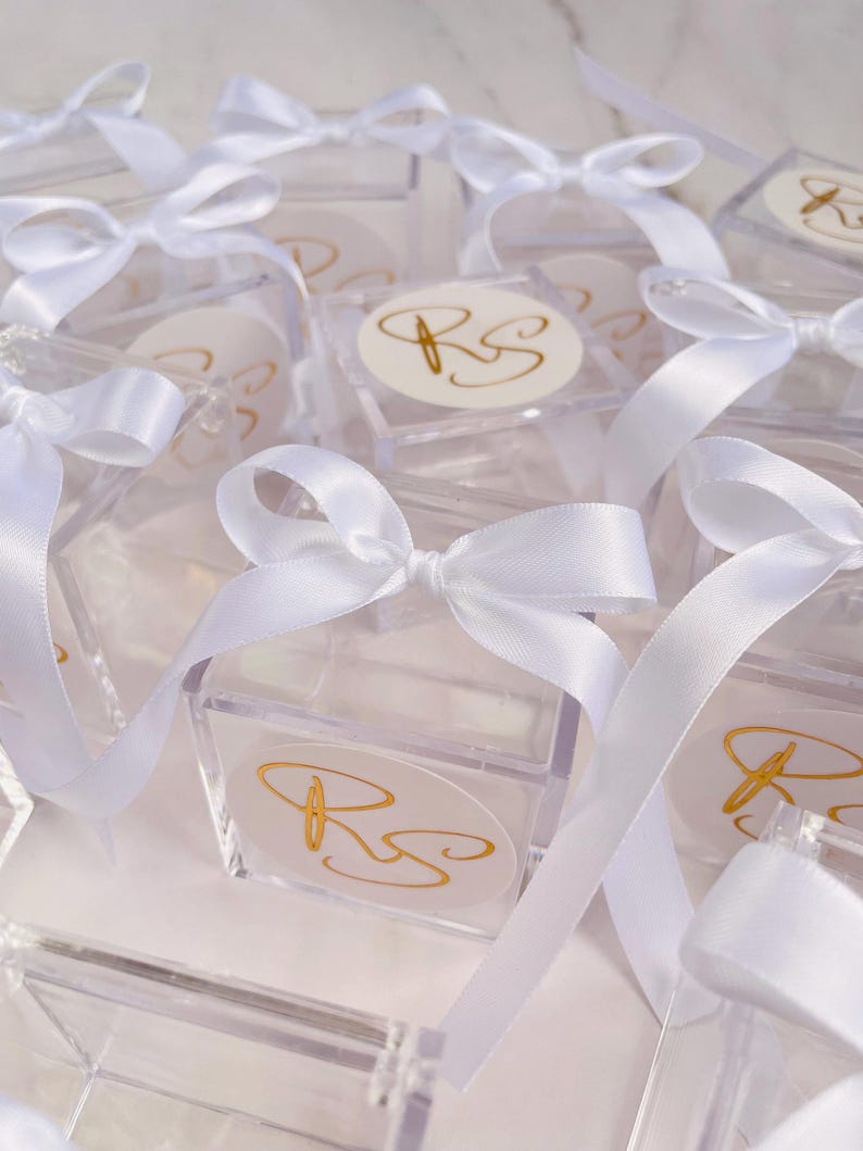 May include: Clear acrylic boxes tied with white satin ribbons and bows. Each box features a gold monogram on a white oval sticker. The boxes are arranged closely together, creating a visually appealing display.