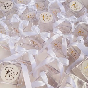 May include: A collection of clear, square boxes tied with white satin ribbons. Each box has a white circular sticker with the gold letters "RS". The boxes are arranged in a scattered pattern, creating a visually appealing display.