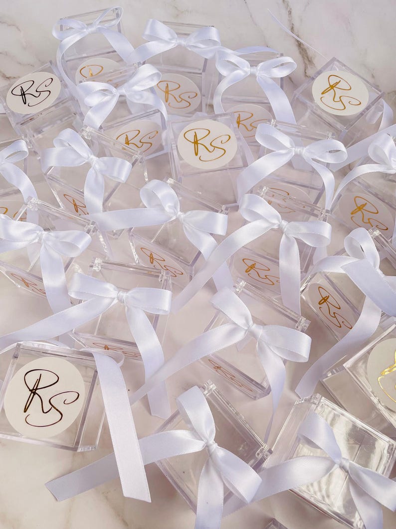May include: A collection of clear, square boxes tied with white satin ribbons. Each box has a white circular sticker with the gold letters "RS". The boxes are arranged in a scattered pattern, creating a visually appealing display.