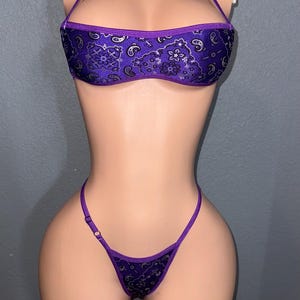 May include: A two-piece purple bikini set with a paisley pattern. The top is a halter style, and the bottom is a thong. The fabric has a shiny appearance.