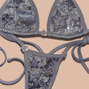 May include: A grey bikini set featuring a triangle top and matching bottoms with tie-side closures. The fabric is adorned with dark mesh star-shaped cutouts, scattered clear rhinestones, and star-shaped rhinestone embellishments on the straps.