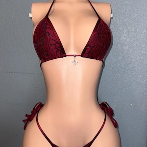 May include: Burgundy bikini set with a triangle top and a thong bottom. The top features a delicate lace pattern and a small silver charm. The bottoms have tie sides. The set is displayed on a mannequin.
