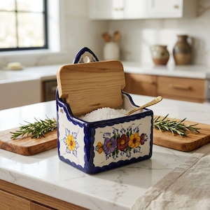 May include: A ceramic salt box with a hinged wooden lid, featuring a white base with blue scalloped edges and hand-painted floral motifs in yellow, purple, and red. The container is filled with white salt and includes a small gold-toned spoon.