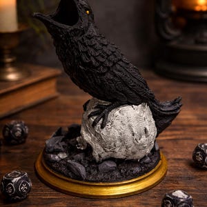 May include: A black raven sculpture with an open beak and yellow eyes, perched on a skull-shaped base. The base is surrounded by dark rocks and sits on a gold-rimmed wooden platform. Decorative dice are scattered around.