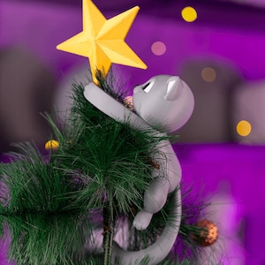 May include: A grey cat Christmas tree topper reaching for a yellow star. The cat is climbing a green artificial Christmas tree. The background is a blurred purple with yellow and orange bokeh lights.