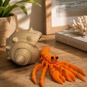 May include: A bright orange articulated hermit crab figurine with black eyes positioned next to a large, beige, spiraled seashell. The crab features multiple segmented legs and claws, showcasing a detailed, textured surface throughout its body.