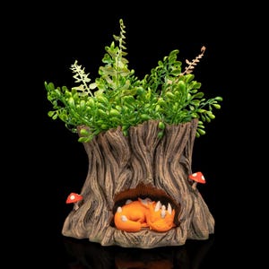 May include: A decorative tree stump planter featuring a sleeping orange fox figurine in a cave. The planter is filled with green foliage and has two red and white mushroom figurines. The planter is made of a brown, textured material, perfect for a small plant.