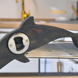 May include: A dolphin-shaped bottle opener made of dark grey wood. It features a silver metal bottle opener mechanism. The dolphin design includes a fin and tail, and it rests on a metal rack.