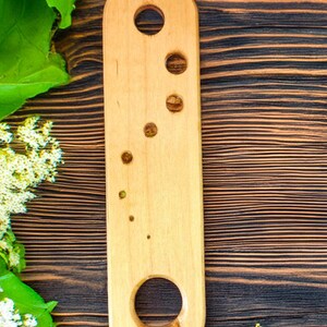 May include: A light-colored wooden bottle opener with a series of circular holes. The opener has a rounded top and bottom, and is set against a dark wood grain background. Green leaves and white flowers are in the corners.