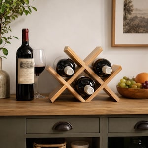 May include: A light-colored wooden wine rack with an X-shaped design, holding three dark glass wine bottles. The rack is constructed from smooth, natural-toned wood slats arranged in a geometric pattern to securely cradle the bottles horizontally.